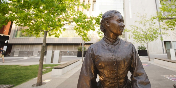 A bronze statue of a woman (Saint Mary MacKillop) stands in a tree-lined courtyard on ACU Melbourne Campus