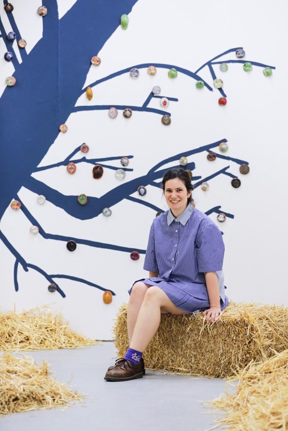 The artist (a woman with dark hair) sits on a hay bale. She wears a blue dress and is smiling