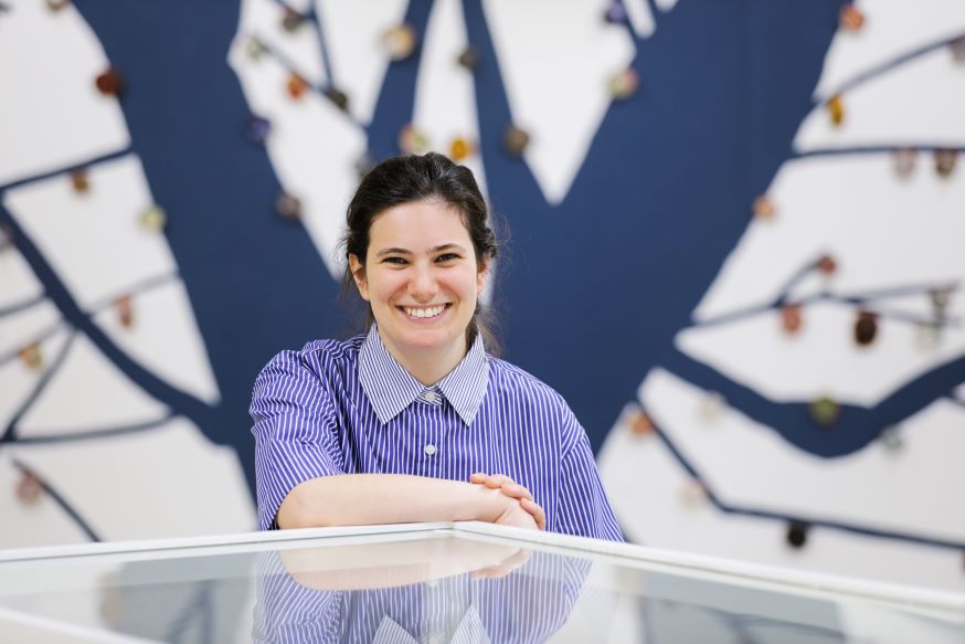 The artist (a woman with dark hair) stands leaning on a display cabinet. She wears a blue dress and is smiling.