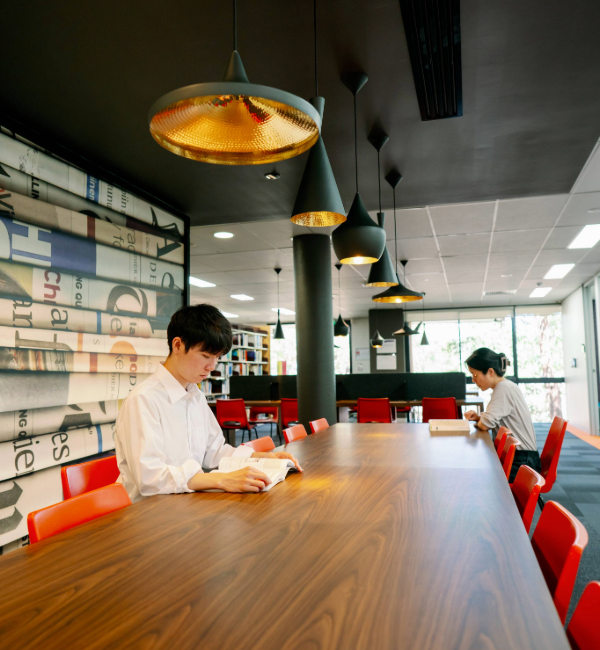 Two people reading books in a library that are sitting on different sides of a long wooden table.