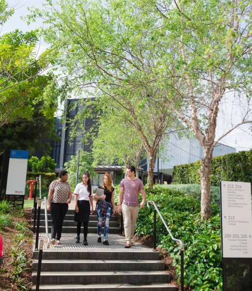 Four university students walk down outdoor steps surrounded by green trees and landscaped gardens on a campus, with modern buildings and directional signage visible in the background.