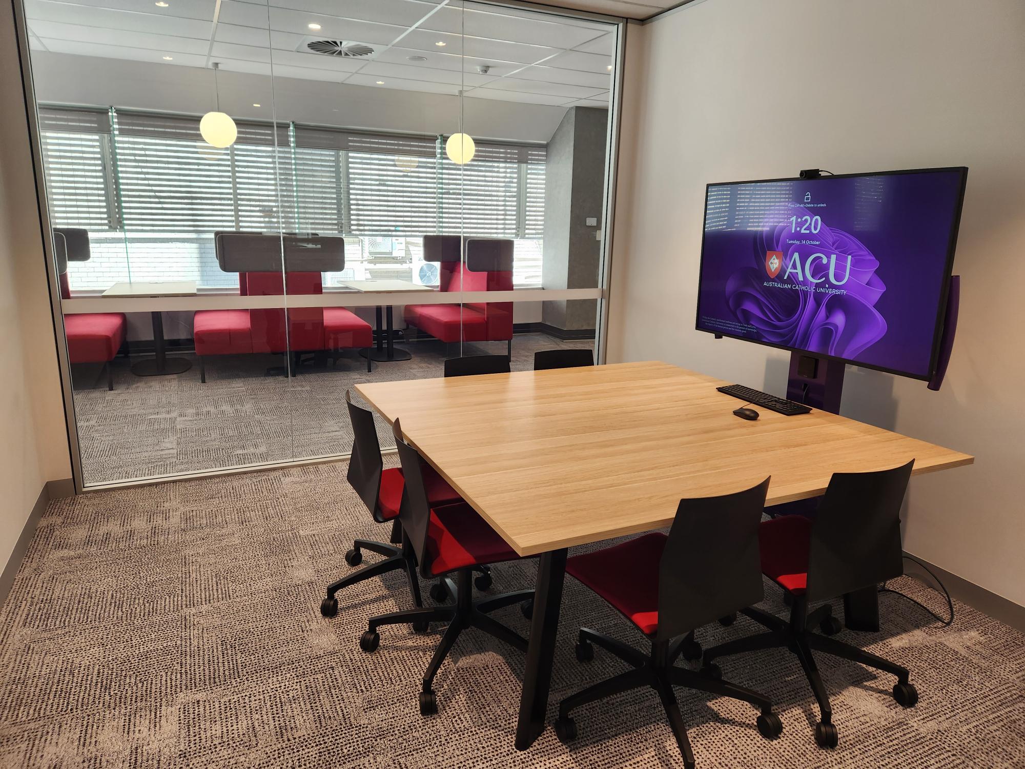 A glass-walled study room with a white table and a large PC screen with the ACU screensaver displaying.